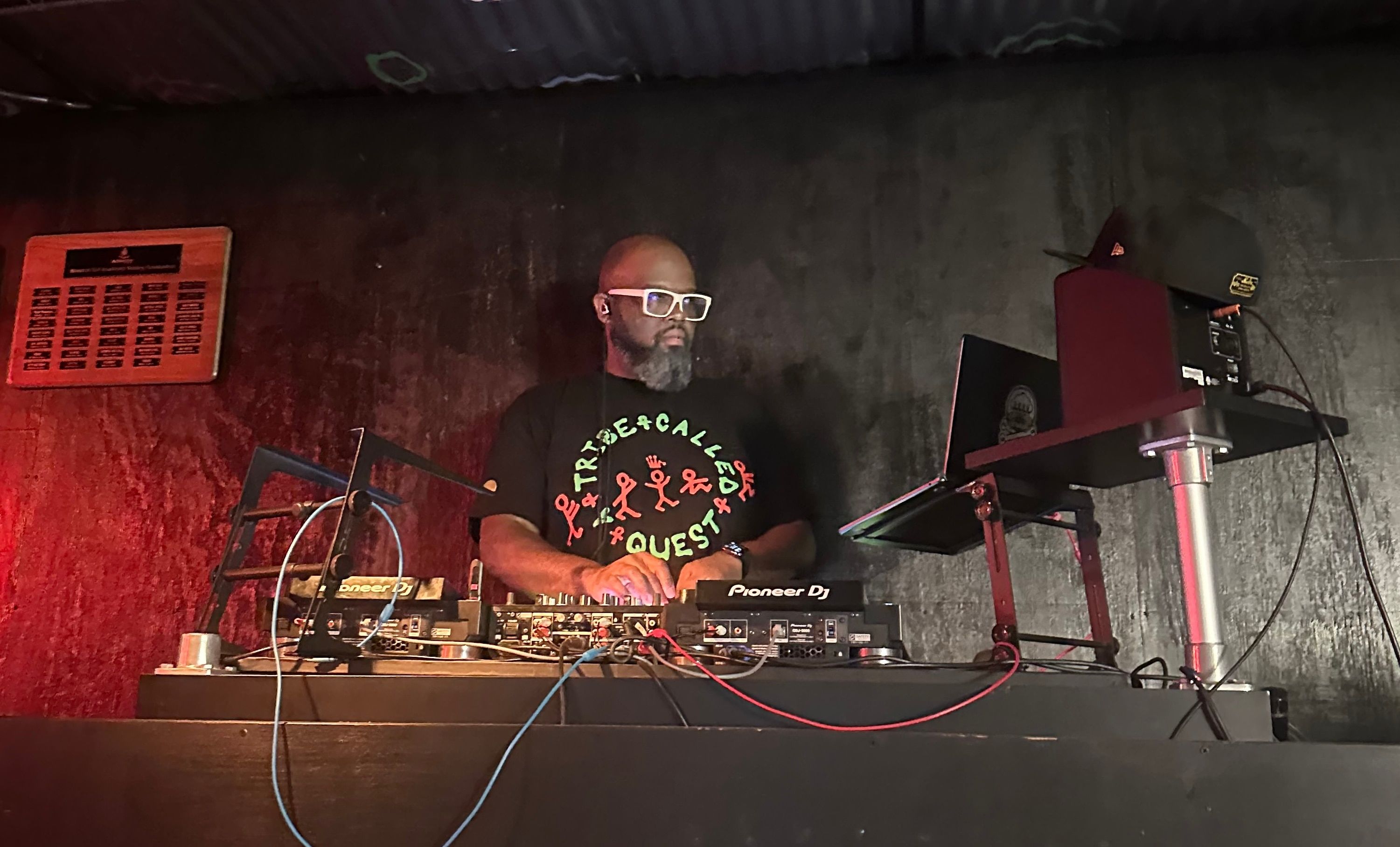 Dwight at the decks in a dark room, hands on the mixer, laptop stand to his right.